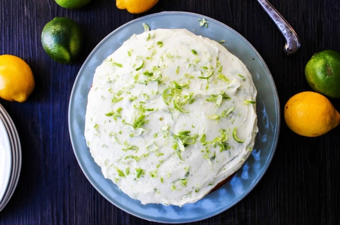 Top down view of Lemon Lime Layer Cake on a blue plate on a table with lemons and limes on the table