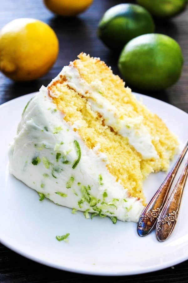 Slice of two layer lemon cake with lemon lime buttercream frosting on a white plate with forks. Lemons and limes on table in background