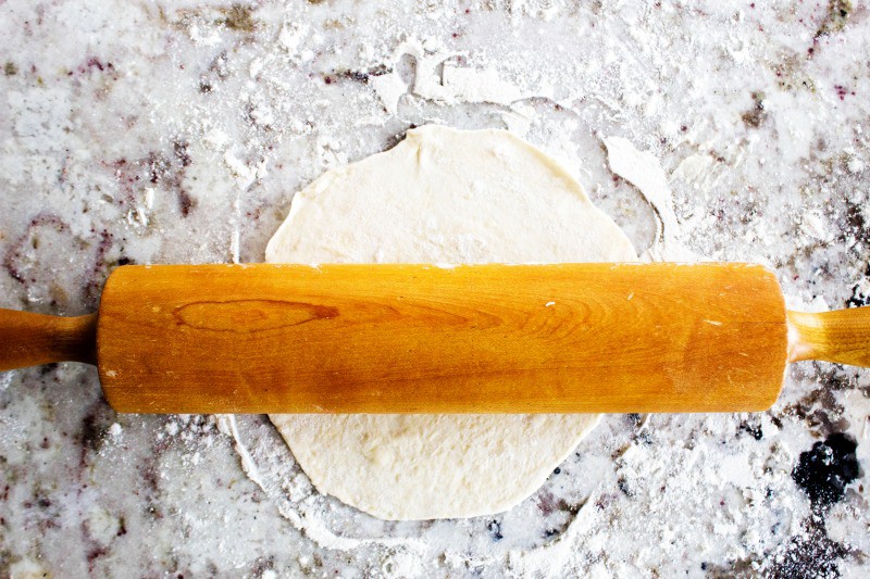 Flour Tortilla dough being rolled out on a floured countertop with a wooden rolling pin