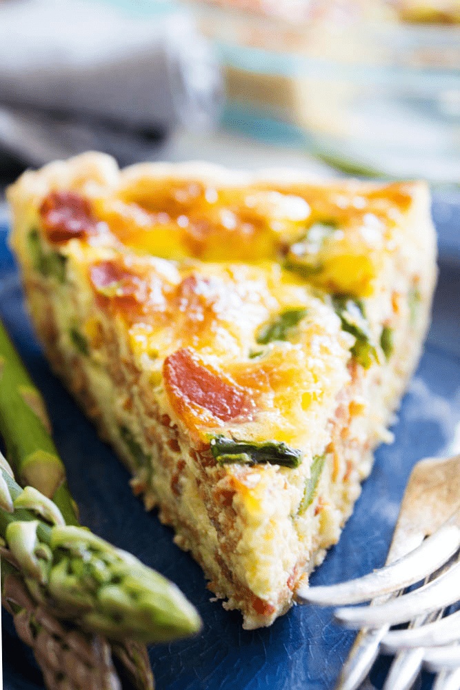 A slice of quiche on a blue plate with forks and asparagus.