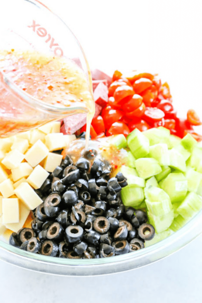 Easy Pasta Salad with Italian dressing, penne pasta, tomatoes, cucumbers, cheese, olives and salami in a bowl not mixed, dressing being poured  into bowl