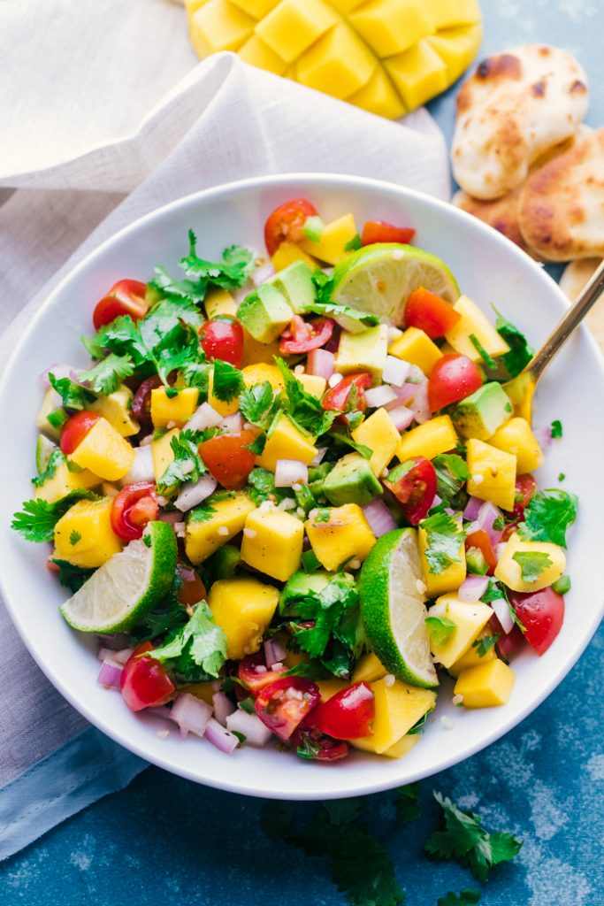 Bowl of Mango Salsa, with lime slices, cilantro, tomato, avocado, and onion in a white bowl with napkin on table