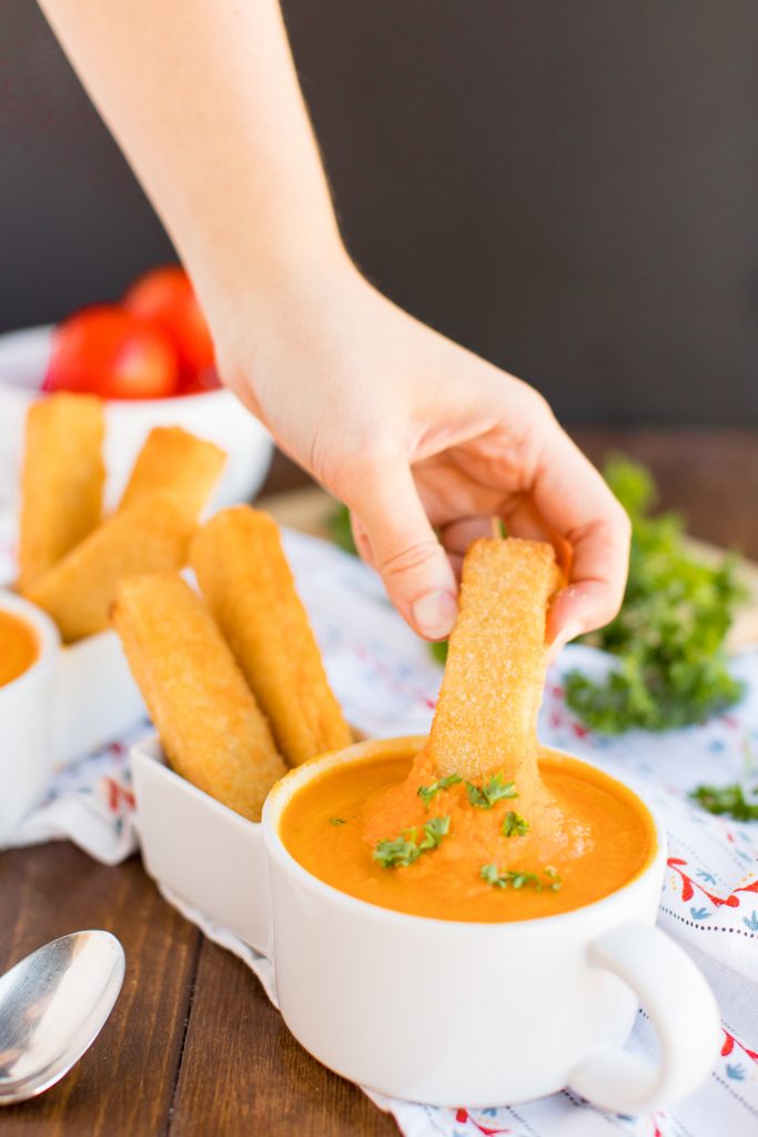 hand dipping a grilled cheese stick in a mug full of tomato soup