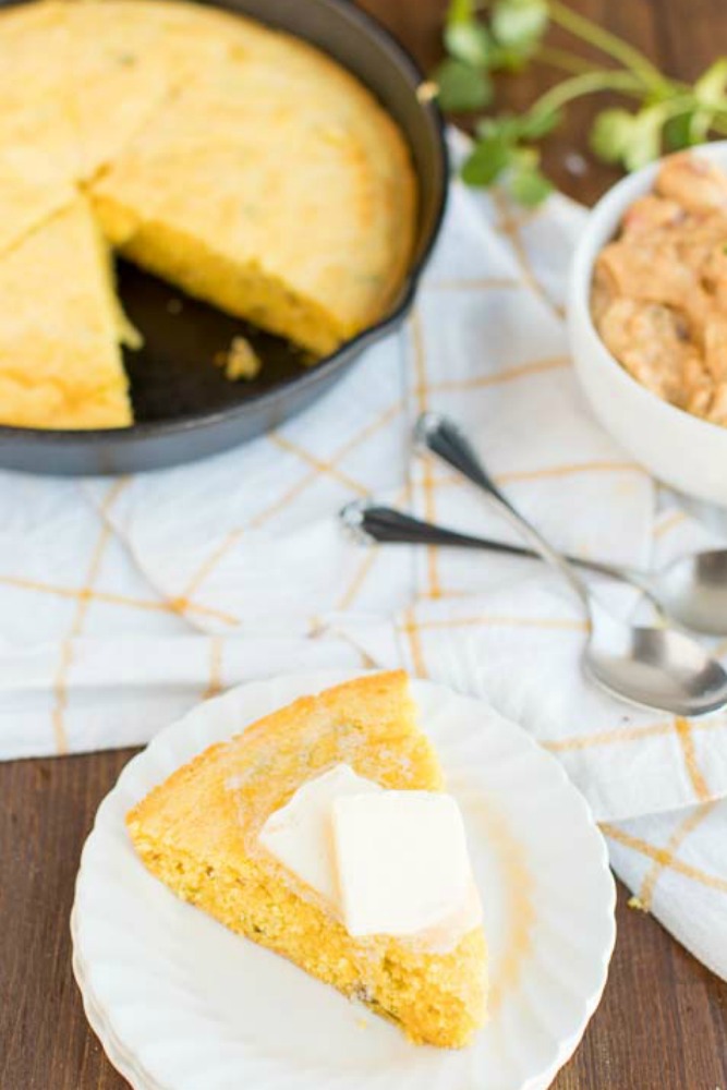 photo of a slice of cornbread on a plate with butter on top, and skillet with cornbread sliced in it