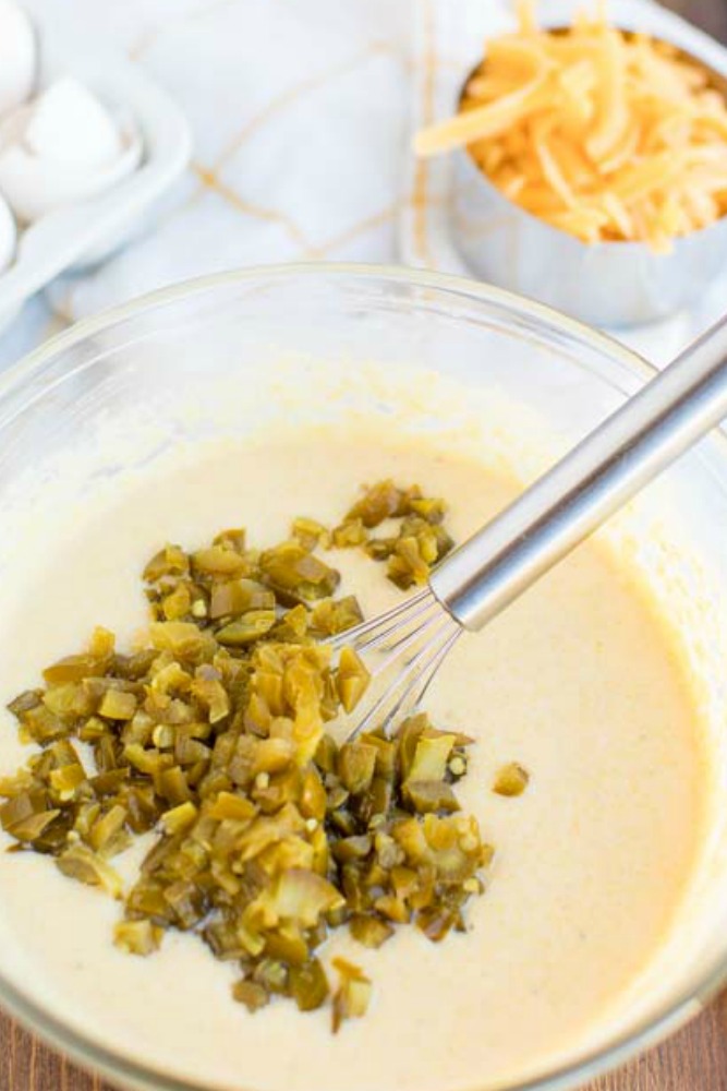 cornbread batter in a glass bowl with jalapenos being stirred in with a metal whisk