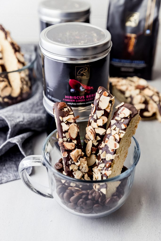 photo showing biscotti in a glass mug with coffee beans at the bottom, with metal jars of private reserve coffee and tea in background