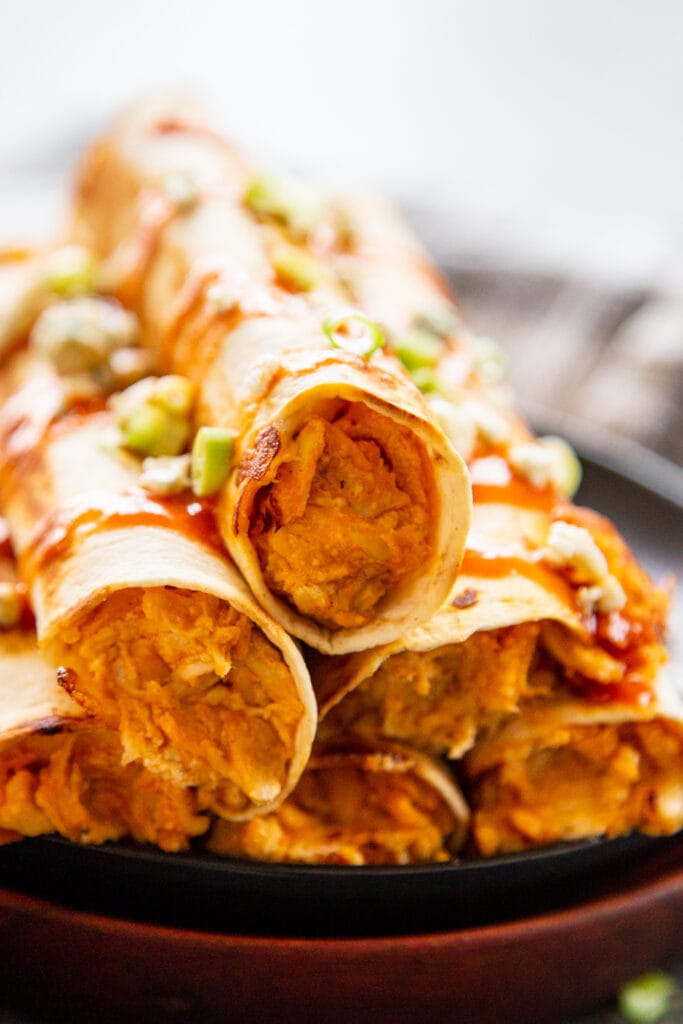 stack of buffalo chicken taquitos on a plate drizzles in sauce and topped with green onions and blue cheese